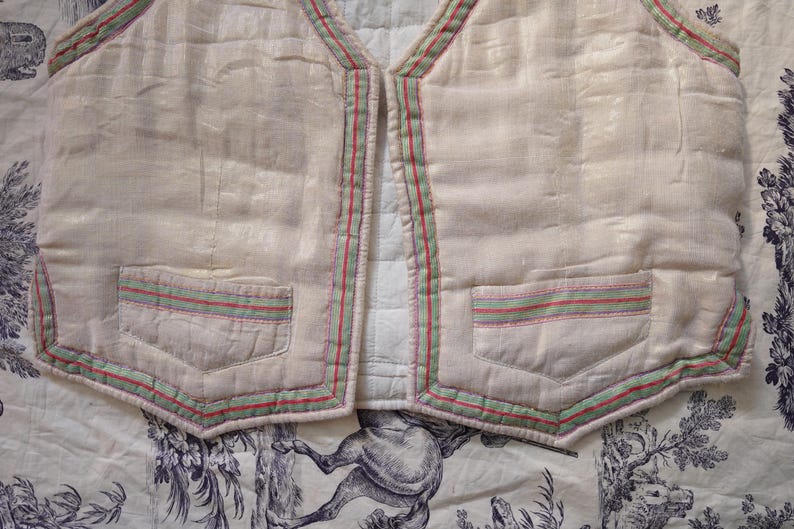 May include: A white quilted vest with green, red, and pink trim. The vest has two pockets on the front and a V-neck.