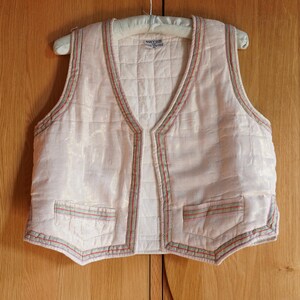 May include: A white quilted vest with a V-neck and a decorative trim of green, red, and white stitching. The vest has two pockets on the front.