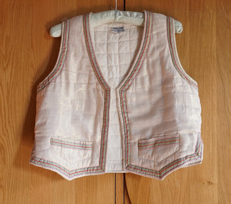 May include: A white quilted vest with a V-neck and a decorative trim of green, red, and white stitching. The vest has two pockets on the front.
