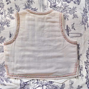 May include: A white sleeveless vest with a red and green trim around the edges. The vest is made of a lightweight fabric and has a simple design.