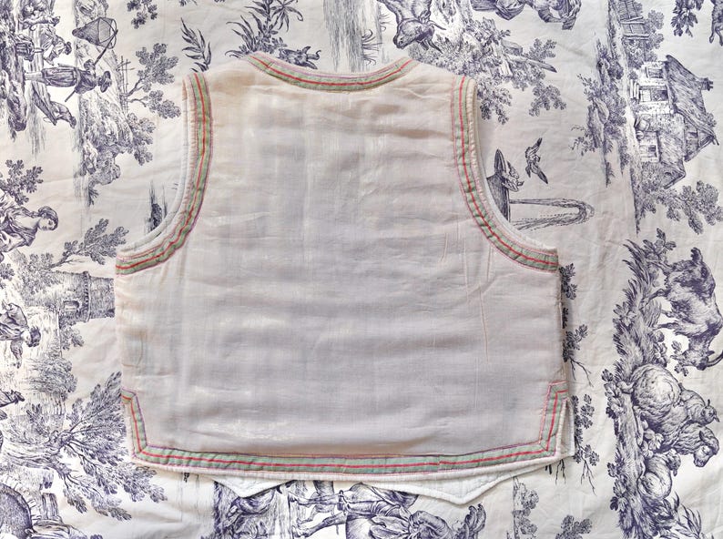 May include: A white sleeveless vest with a red and green trim around the edges. The vest is made of a lightweight fabric and has a simple design.
