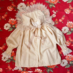 Könnte beinhalten: Cremefarbene Vintage-Bluse mit großem Spitzenkragen und Manschetten. Die Bluse hat lange, gepuffte Ärmel und eine Satinbandschleife am Ausschnitt. Der Stoff hat ein leicht gealtertes Aussehen, was auf seinen Vintage-Charakter hindeutet. Die Bluse ist vorne geknöpft.