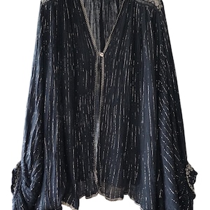 Puede incluir: Top negro de manga larga con rayas plateadas verticales y cuello en V. El top presenta un ribete plateado decorativo a lo largo del escote y los puños. Una cadena plateada con cierre de botón adorna el frente.