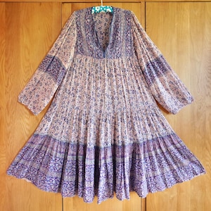 Könnte beinhalten: Ein langärmliges Kleid mit V-Ausschnitt und Stufenrock. Das Kleid hat ein Paisley-Muster in Lila- und Rosatönen. Das Kleid hängt an einem Kleiderbügel vor einem hölzernen Hintergrund.