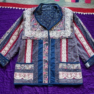 May include: A blue quilted jacket with a patchwork design featuring floral prints and white lace trim. The jacket has a collar, long sleeves, and a button closure.