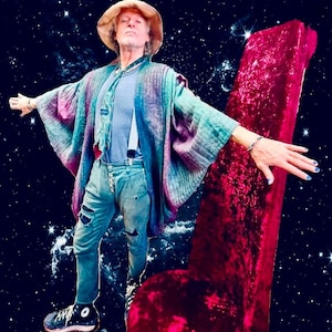 May include: A man wearing a blue and green patterned robe, blue jeans, and a brown hat stands in front of a large, red velvet chair. The background is a dark blue with white stars.
