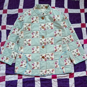 May include: A vintage quilted jacket with a floral and plaid pattern in shades of blue, white, and pink. The jacket has a mandarin collar, a button-up front, and two patch pockets.