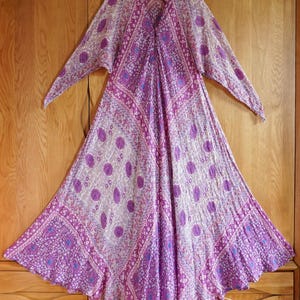 Puede incluir: Un vestido largo y fluido con un vibrante estampado morado y blanco. El vestido presenta un cuello en V, mangas largas y una falda acampanada. La tela está adornada con diseños geométricos y de lunares, creando una estética bohemia.