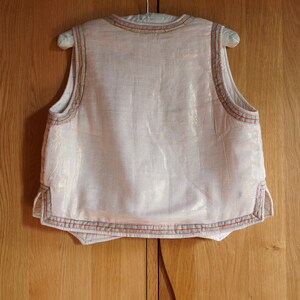 May include: A sleeveless white vest with a light pink hue, featuring a decorative trim of red, green, and beige along the edges.