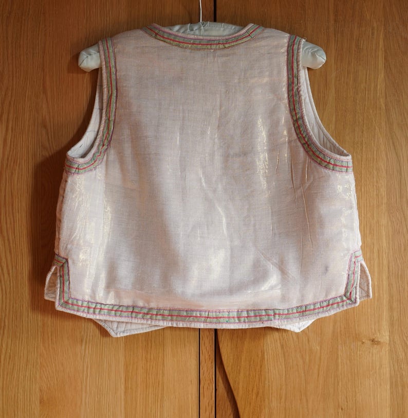 May include: A sleeveless white vest with a light pink hue, featuring a decorative trim of red, green, and beige along the edges.