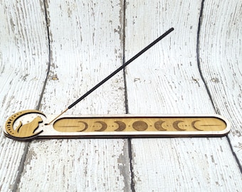 Puede incluir: Quemador de incienso de madera con un diseño de gato y un diseño de fase lunar. El quemador de incienso está hecho de madera marrón claro y tiene un palo de incienso negro descansando sobre él.