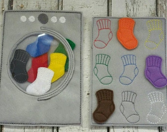 May include: A felt toy washing machine with a clear window showing colorful socks inside.  The machine has a control panel with buttons and a dial.  There are nine felt socks on a separate panel, some with matching colors to the socks in the machine.