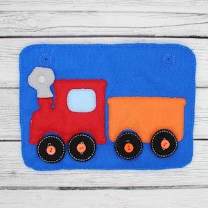 May include: A blue felt train-themed wall hanging. The train is red with a light blue window, and the attached car is orange. The wheels are black with orange button centers. A gray smoke stack is attached to the train.