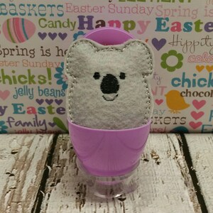 May include: A grey felt koala bear toy inside a purple plastic Easter egg. The egg is sitting on a wooden surface. The background is a white and pink patterned paper with the text "Candy Happy Easter" and "Hippity colo".