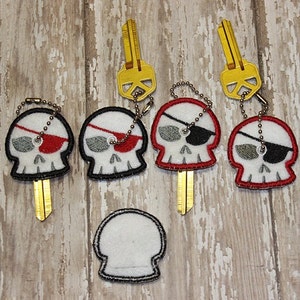 Puede incluir: Cinco llaveros con un diseño de calavera con parche en el ojo. Las calaveras están bordadas en negro, blanco y rojo sobre una tela blanca. Los llaveros están unidos a llaves de color dorado.