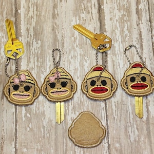 May include: Four keychains featuring embroidered monkey faces with different expressions. Two monkeys wear pink bows and sunglasses, one wears a red hat, and one wears a red hat and has a red mouth. The keychains are attached to gold keys.