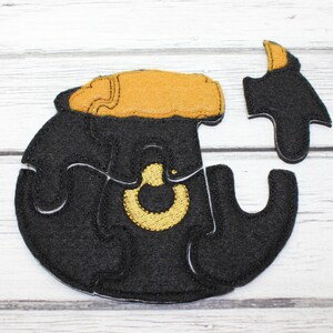 Puede incluir: Un rompecabezas de fieltro negro de un caldero con un borde y un asa dorados. El rompecabezas está formado por cinco piezas.