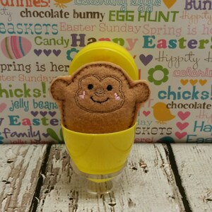 May include: A brown plush monkey toy with a happy face sits inside a yellow plastic egg. The egg is on a white wooden surface with a background of colorful Easter-themed text.