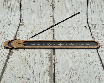 Puede incluir: Un porta incienso de madera con un diseño negro y dorado que presenta una luna creciente y estrellas. El porta incienso está diseñado para sostener un solo palo de incienso.