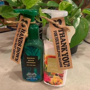 May include: Two bottles of foaming hand soap with brown paper gift tags. The left bottle is green and says "Waikiki Beach Coconut" and "Hands Down You're the Best Around!" The right bottle is white with floral designs and says "Bright Bloc" and "Thank You! For Your Helping Hands".