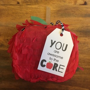 Könnte beinhalten: Eine rote, apfelförmige Piñata mit einem weißen Etikett, auf dem "YOU are awesome to the CORE" mit einem roten Apfel-Motiv steht.