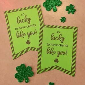 Puede incluir: Dos tarjetas rayadas de color verde y marrón con el texto "SO lucky to have clients like you!" impreso en ellas. Las tarjetas están decoradas con tréboles de purpurina verde.
