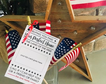 May include: A white card with black text that reads "Helping you find your perfect home. The land of the free." with a space for a business card. There are three small American flags in a glass vase. The card is tied with a red ribbon.