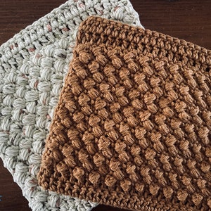May include: Two crocheted dishcloths, one in a light beige colour and the other in a brown colour. Both dishcloths have a textured, puffy design.