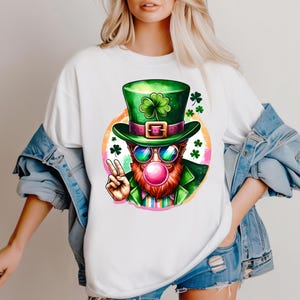 Puede incluir: Camiseta blanca con una ilustración de dibujos animados de un duende con un sombrero verde, gafas de sol y soplando una burbuja rosa. El duende está levantando dos dedos en señal de paz.