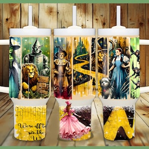 May include: Three white tumblers with a yellow brick road, a wizard, a lion, a tin man, and Dorothy from the Wizard of Oz. The tumblers have a wood grain background and the text "We're off to see the Wizard".