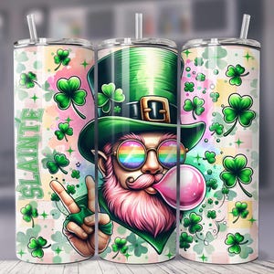 May include: A stainless steel tumbler with a green, pink, and white design. The design features a cartoon leprechaun wearing a green hat, rainbow sunglasses, and blowing a bubble. The text "Slainte" is written on the tumbler.
