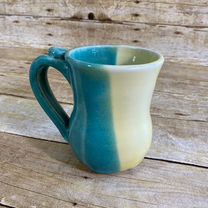 May include: Handmade ceramic mug with a teal and cream color scheme. The mug features a curved handle and a unique design, perfect for enjoying your favorite beverage. The mug is approximately 4 inches tall.