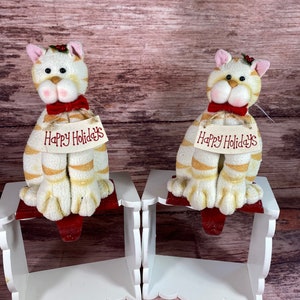 May include: Two white and gold striped cat stocking holders with red bows and the text "Happy Holidays" on a white shelf with scalloped edges.