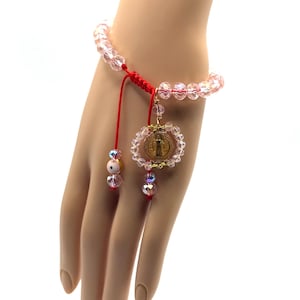 May include: A pink beaded bracelet with a red cord and a gold medallion. The bracelet features clear, faceted beads and a dangling charm with a religious image. Additional pink and iridescent beads adorn the red cord.