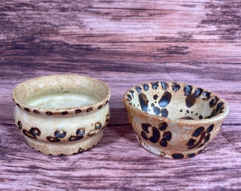 May include: Two small ceramic bowls with a brown and white speckled glaze. The bowls have a rounded shape and a slightly flared rim.