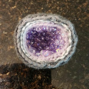 May include: A circular crocheted geode-inspired object featuring a dark grey outer border, a white middle ring, and a light purple inner core filled with numerous small, faceted, translucent purple beads that mimic the appearance of crystals.