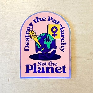 Peut inclure: Un autocollant rose avec le texte "Destroy the Patriarchy Not the Planet". L'autocollant représente un personnage de la Terre de dessin animé tenant une massue à pointes et un panneau avec le symbole féminin. La Terre est bleue et verte, avec un visage souriant.