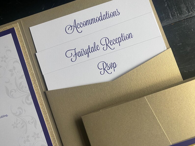 May include: A gold wedding invitation suite with inserts. The inserts have the words "Accommodations", "Fairytale Reception", and "RSVP" in a dark blue script font. The invitation has a purple and gold border.