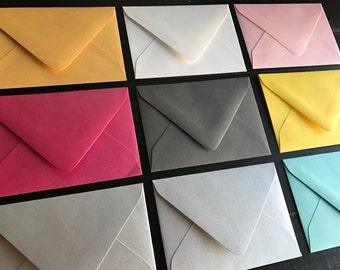 Puede incluir: Una colección de diez sobres de varios colores, incluyendo amarillo, blanco, rosa, gris, rojo, azul claro y crema. Los sobres están dispuestos en tres filas, con tres sobres en cada fila.