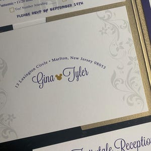 May include: Wedding invitation with a white background and a gold border. The invitation features the names "Gina & Tyler" in a purple script font, with a gold Mickey Mouse head. The address is printed in a curved font.