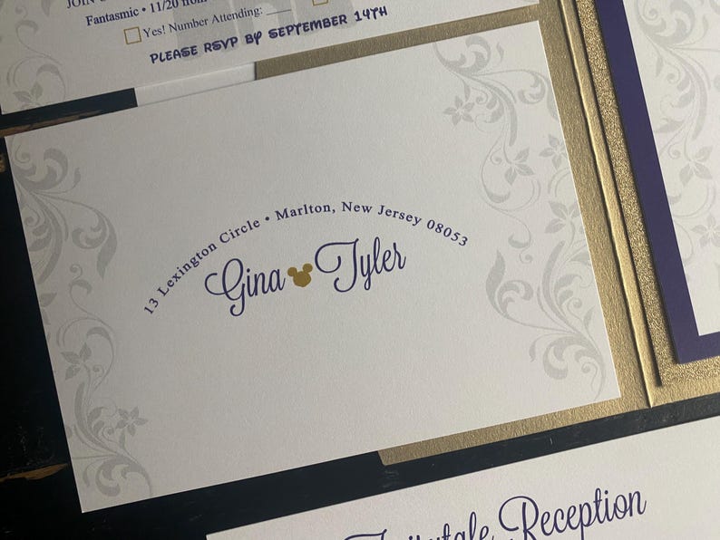 May include: Wedding invitation with a white background and a gold border. The invitation features the names "Gina & Tyler" in a purple script font, with a gold Mickey Mouse head. The address is printed in a curved font.