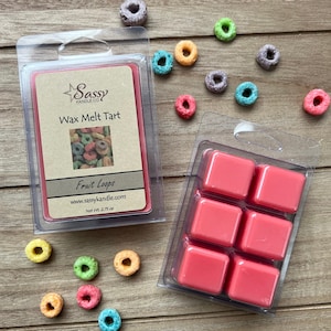 May include: A wax melt tart in a clear plastic container with a red wax melt inside. The wax melt is labeled "Sassy Candle Co. Wax Melt Tart Fruit Loops" and has a picture of Fruit Loops cereal on the label. The container is sitting on a wooden surface with colorful cereal rings scattered around it.