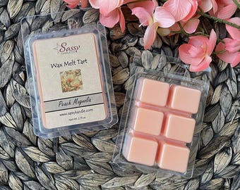 May include: A six-pack of wax melt tarts in a clear plastic container. The wax melts are a light peach color. The label on the container reads "Sassy Candle Co. Wax Melt Tart Peach Magnolia www.sassycandle.com Net Wt. 2.75 oz."