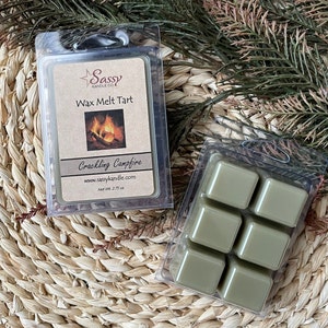 May include: A six-pack wax melt tart in a clear plastic container. The wax melts are a sage green color. The label on the container reads "Sassy Candle Co. Wax Melt Tart Crackling Campfire www.sassycandle.com Net Wt. 2.25 oz."