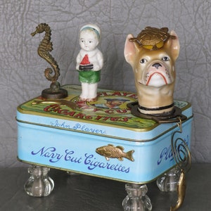 May include: A blue and white tin box with the words "Navy Cut Cigarettes" and "John Player's" printed on it. The box is decorated with a gold fish and has four glass feet. A porcelain figurine of a dog wearing a turtle on its head sits on top of the box. A porcelain figurine of a child holding a boat sits on the left side of the box. A gold seahorse figurine sits on the right side of the box.