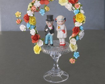 Wedding or Anniversary cake topper.  Bisque 1920s bride and groom set atop cut crystal base adorned with an arch of colorful paper flowers.