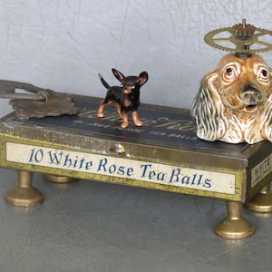 May include: A vintage tin box with a gold base and legs, featuring a blue and white label that reads "10 White Rose Tea Balls". A porcelain dog figurine with a gear-shaped hat sits on top of the box, and a small black and brown chihuahua figurine stands on the box's surface.