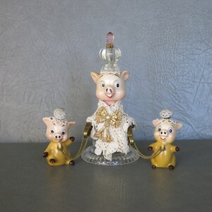 May include: Three ceramic pig figurines, one larger than the other two. The larger pig is wearing a white lace dress with a gold bow and a clear glass crown with a pink top. The two smaller pigs are wearing hats and are connected to the larger pig by gold chains.