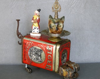 Assemblage circus cat.  Porcelain cat head, Tiger tobacco tin, brass lion's paw castors, cast iron hat hook, clown, brass dish and finial.