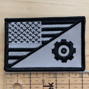 May include: A gray and black embroidered patch featuring a stylized American flag with a gear in the bottom right corner. The gear has a black circle in the center.
